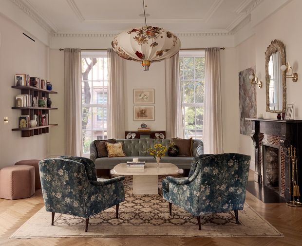 Elegant, traditionally detailed living room with a symmetrical seating arrangement, ornate ceiling mouldings, tall sash windows dressed in sheer curtains, a decorative floral pendant light, fireplace with dark surround and carved mirror, wall shelving, and a patterned area rug. The palette is warm and refined with muted floral upholstery, cream walls, timber flooring, and vintage-inspired furnishings.