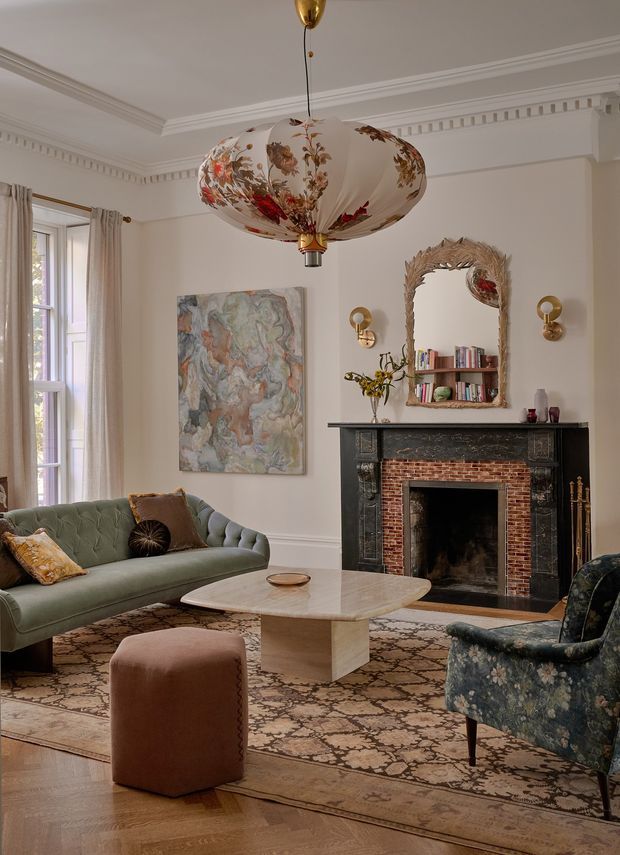 Elegant traditional living room with high ceilings, ornate crown moulding and dentil detailing, a decorative floral pendant light, tall sash window with curtains, tufted sage-green sofa, patterned armchair, sculptural stone coffee table, and a restored fireplace with black mantel and exposed brick surround. A wall mirror above the mantel reflects part of the room, while an artwork, books, brass wall sconces, and layered rug add refined character.