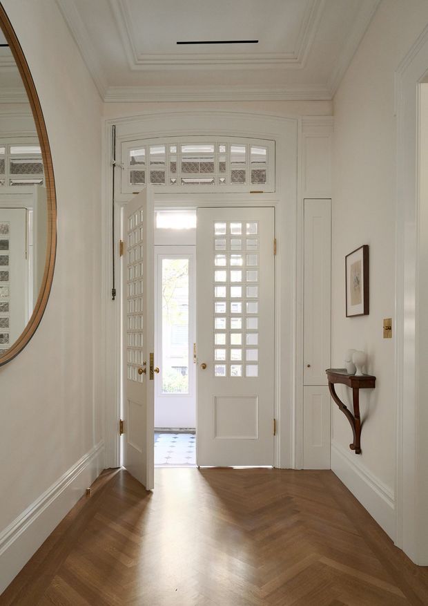 Bright, elegant residential entry hall with a white paneled door and fanlight glazing, ornate ceiling mouldings, tall decorative wall panelling, a large round timber-framed mirror, and a slim dark timber console table. The floor is finished in a warm herringbone timber pattern, and the open doorway leads to a lighter tiled space beyond.