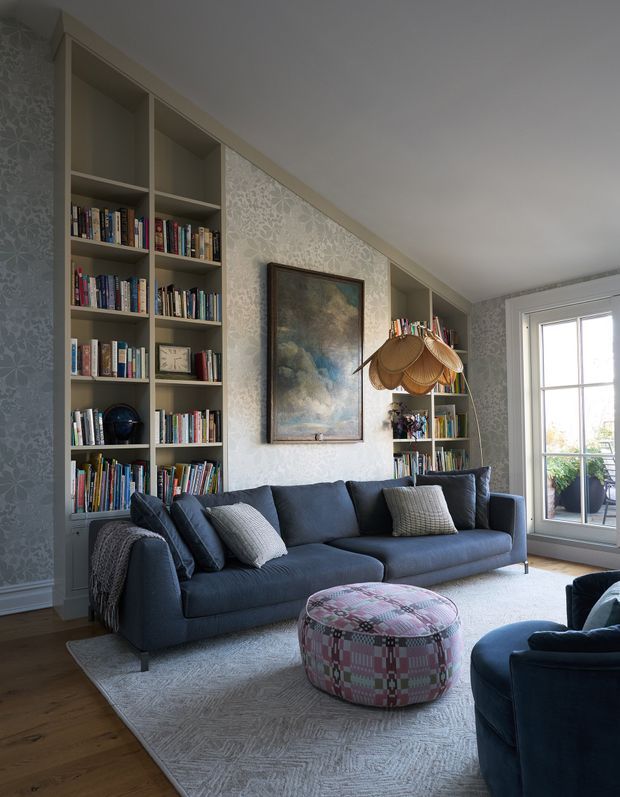 A cosy living room with a deep navy sectional sofa, matching armchair, patterned ottoman, and light textured rug. Full-height built-in bookshelves flank a framed artwork, while delicate patterned wallpaper, dark timber flooring, and a woven pendant light create a layered, eclectic interior. A glazed exterior door or tall window opens to a small balcony or terrace.