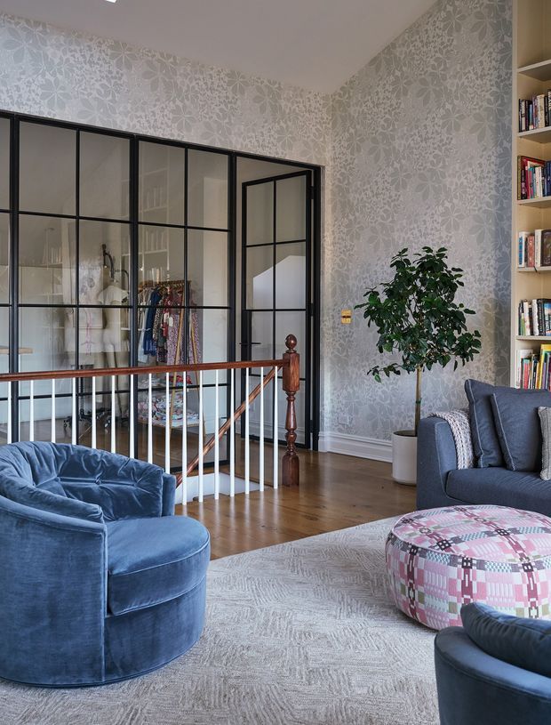 A stylish living room or sitting area with blue velvet lounge chairs and sofa, a large pale patterned rug, and a colourful patchwork ottoman. The room features ornate light grey floral wallpaper, warm timber flooring, a traditional timber stair balustrade, black-framed glass doors enclosing a wardrobe or dressing area, and built-in open bookshelves. The space blends classic decorative finishes with contemporary steel-framed glazing and eclectic furnishings.