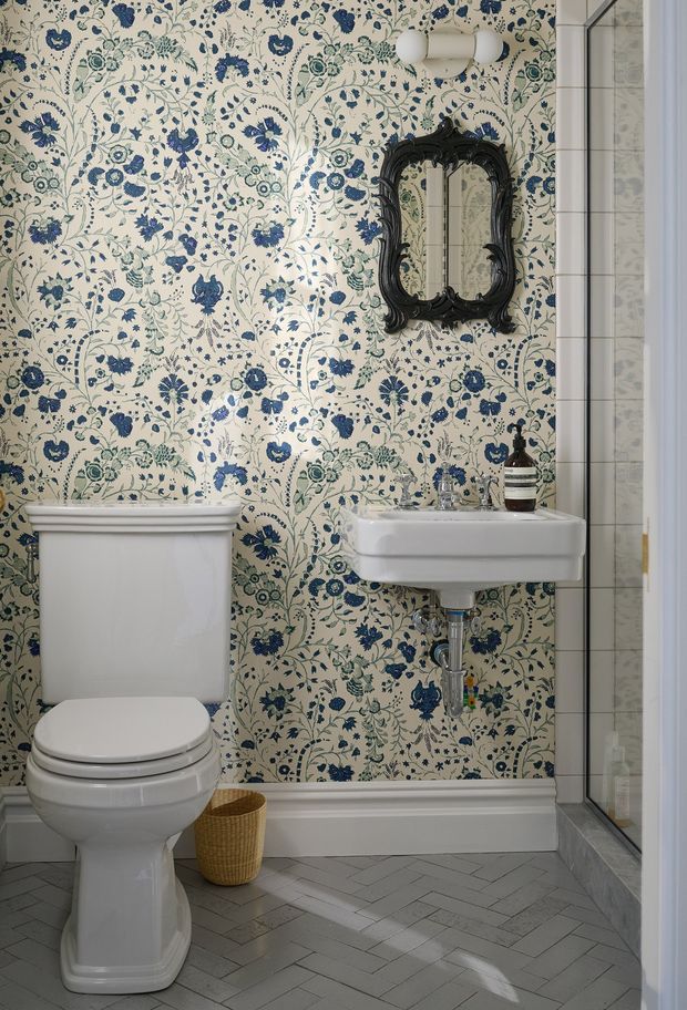 A compact, highly decorative bathroom featuring a white pedestal-style wall-mounted basin and toilet against blue-and-cream botanical floral wallpaper. A dark ornate framed mirror and twin globe wall light add traditional character, while grey herringbone floor tiles and a glass shower enclosure complete the space.