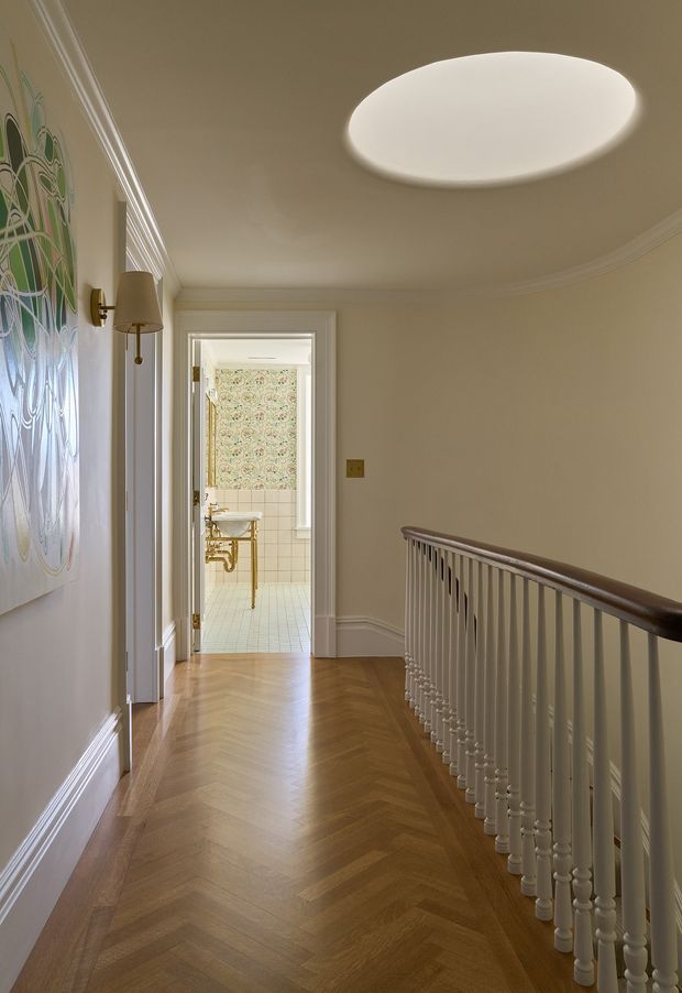 Elegant upper-floor hallway or landing with a warm herringbone timber floor, cream painted walls, ornate cornices and skirting, and a decorative white balustrade with dark timber handrail. A large patterned glass or artwork panel and wall sconce line the left wall, while a sculptural oval ceiling light illuminates the space. An open doorway reveals a bathroom with patterned wallpaper, tiled lower walls, and a traditional basin.
