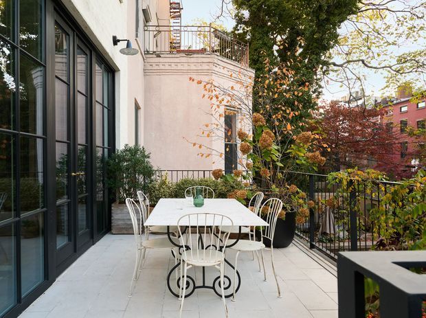 A furnished outdoor terrace overlooking a leafy urban setting, with a white dining table and metal chairs on pale large-format paving. The terrace is enclosed by black metal railings and bordered by planting, while the adjacent building has tall black-framed glazed doors, pale rendered walls, and a small upper balcony with decorative metal balustrading. The scene has an elegant, relaxed contemporary urban character in autumn colours.