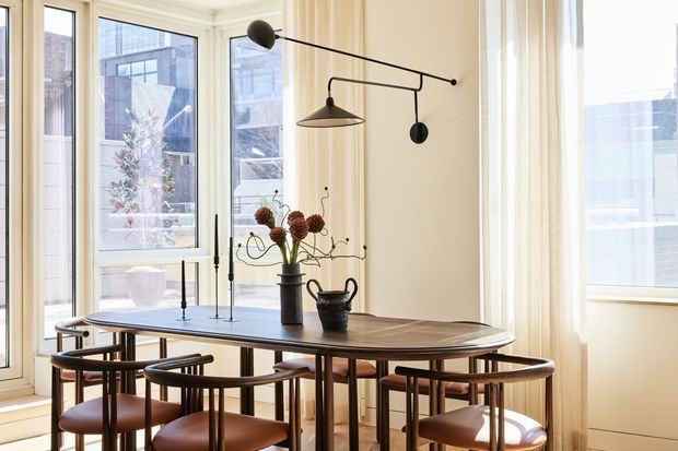 A bright, refined dining area with a large oval timber dining table surrounded by sculptural wooden chairs with brown leather seats. Tall cream-framed windows and sheer curtains fill the room with natural light, while a minimalist black articulated pendant light and floral arrangement provide focal points. Urban buildings are visible through the glazing.