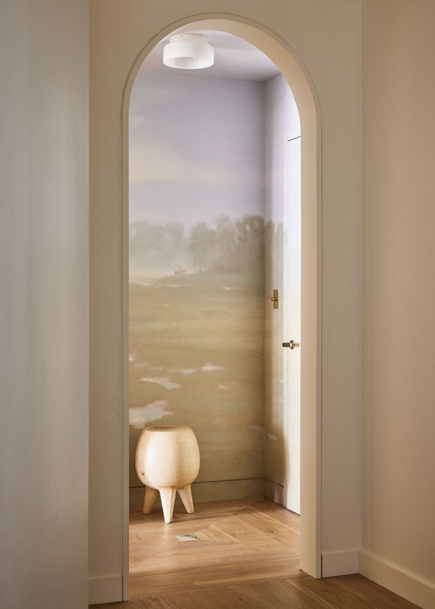 A softly lit interior view through a tall arched doorway into a small, minimalist room. The room features warm timber flooring, pale plaster walls, a white sculptural pendant light, a partially visible white door, and a small rounded stool or side table. The rear wall has a muted atmospheric landscape-style finish or mural in soft beige and green tones.