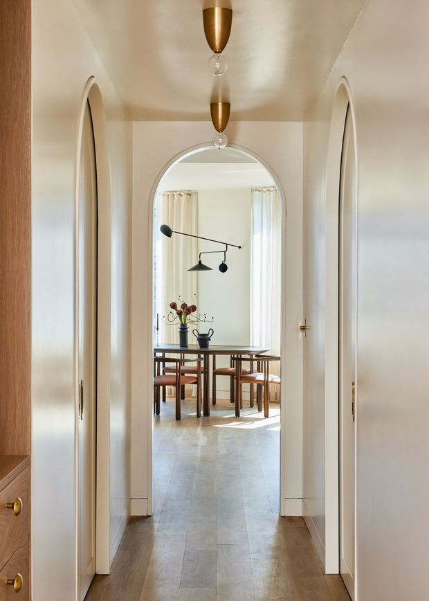 A refined residential hallway with a central arched opening leading into a bright dining room. The corridor has pale cream walls, arched built-in doorways, warm timber flooring, concealed storage, and paired brass pendant lights. The dining room beyond features a timber dining table and chairs, light curtains, and a sculptural black wall light.