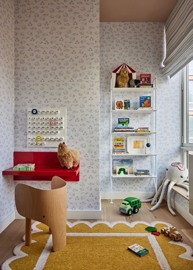 A playful children's room with whimsical animal-patterned wallpaper, a red wall-mounted desk and shelf, children's books and toys, a curved wooden chair, plush octopus, and a mustard-yellow rug with white scalloped lines. Natural light enters through the window with a striped Roman blind.