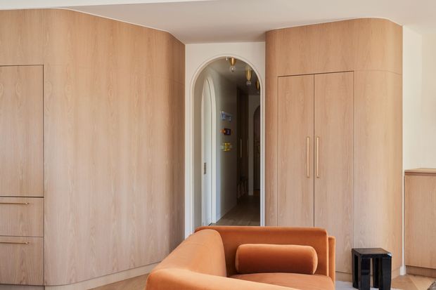 A warm, refined interior featuring extensive curved light timber built-in cabinetry and wardrobes, an arched doorway leading to a hallway, and a sculptural burnt-orange upholstered sofa in the foreground. The room has a smooth light ceiling, pale walls, timber flooring, and restrained contemporary detailing.