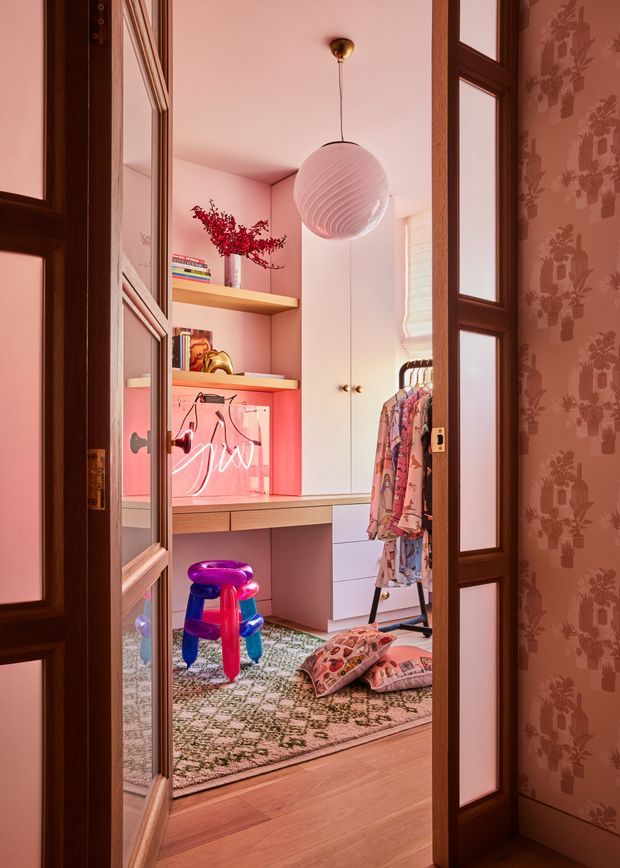A richly styled dressing room or walk-in wardrobe viewed through partially open timber-and-glass panelled doors. The room features pink walls and cabinetry, open shelving with books and accessories, a built-in desk or dressing counter, hanging patterned clothing, a sculptural pendant light, patterned rug, colourful decorative stool, and cushions. Dark timber-framed doors and patterned wallpaper frame the entrance.