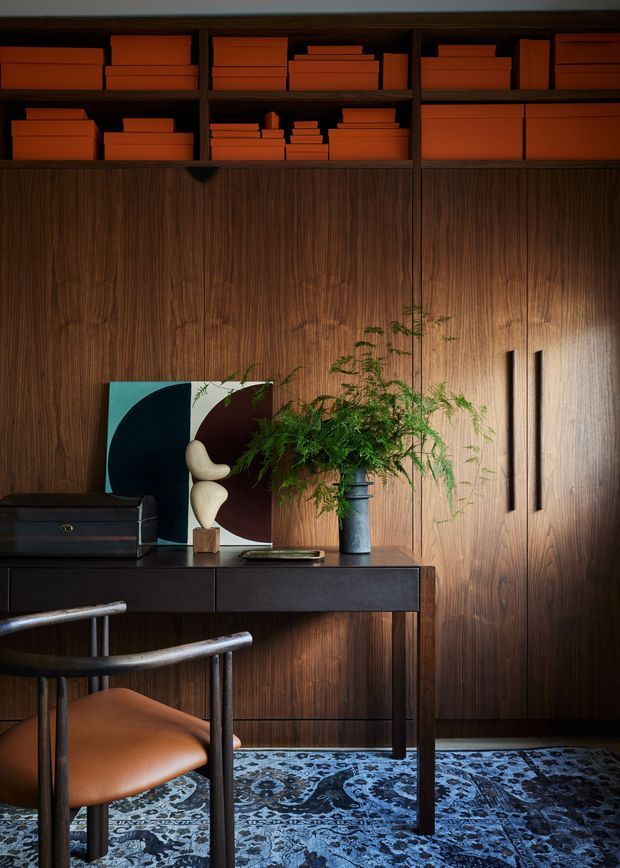 A richly styled study or home office featuring full-height dark timber wall panelling and integrated cabinetry with open shelving above. A slim dark desk holds artwork, a sculptural object, a black storage box and a tall vase of greenery. A leather-and-metal armchair sits on a patterned blue rug, creating a warm, moody and refined atmosphere.