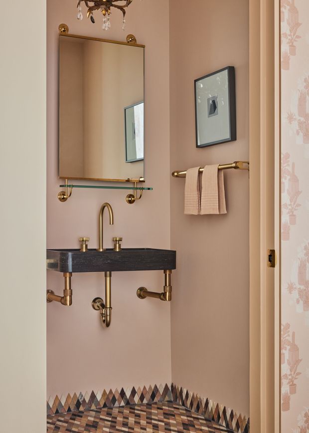 A compact, highly detailed powder room with dusty pink walls, a wall-mounted dark stone basin, exposed brass plumbing and faucets, a large framed mirror with glass shelf, brass towel rail, framed artwork, ornate chandelier, and a striking geometric multicoloured mosaic floor with matching wall border. Decorative patterned wallpaper is visible at the doorway.