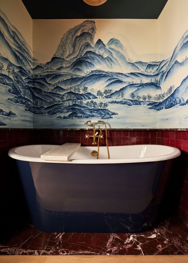 A dramatic bathroom centred on a freestanding deep navy bathtub with a white interior and brass wall-mounted tapware. The walls feature glossy deep-red tiles and an expansive blue-and-white mountain landscape mural, while the floor is finished in richly veined dark red marble. A dark ceiling and warm lighting add contrast.