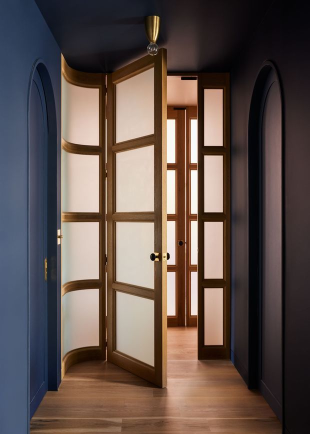 A dramatic interior hallway or entry passage with dark navy-blue walls and ceiling, warm timber flooring, and a series of distinctive timber-framed doors with frosted or translucent panels. The foreground door is open, revealing a brighter adjoining space with matching glazed timber partitions. Arched wall niches or openings frame both sides, while a small brass pendant light adds a warm metallic accent. The composition is refined, moody, and highly architectural.