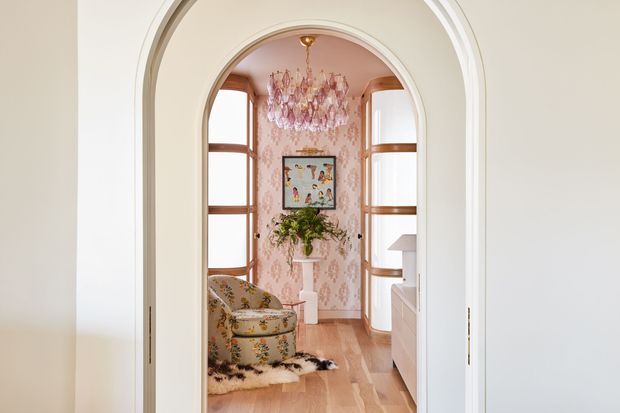 A view through a white arched opening into a richly decorated sitting room or reading nook. The room features curved timber-framed built-in shelving or cabinetry, warm timber flooring, pink patterned wallpaper, a floral upholstered chair, a crystal chandelier, framed artwork, and a central pedestal with greenery. The palette is soft and warm with ornate, playful detailing.