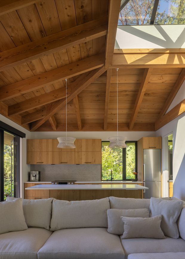 A warm open-plan living and kitchen space viewed from behind a large light-colored sectional sofa. The kitchen features flat-panel timber cabinetry, a long island with a pale benchtop, integrated appliances, and two sculptural pendant lights. Above, a prominent exposed timber post-and-beam ceiling with timber boards and a high clerestory-style glazed opening creates a dramatic vaulted volume. Large dark-framed windows provide abundant natural light and views to trees outside.