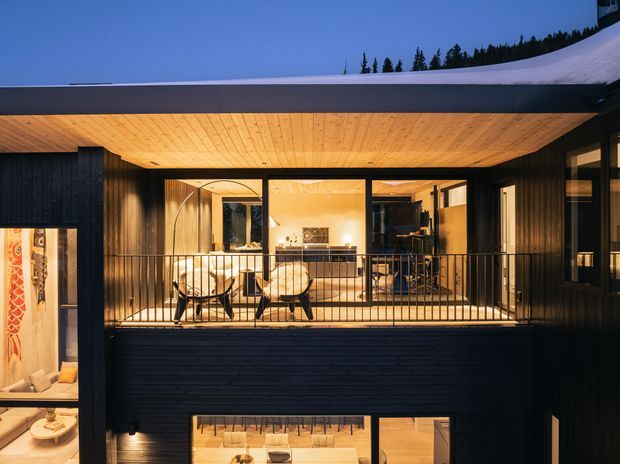 Exterior view of a dark-clad, multi-level modern house in a snowy alpine setting. The image focuses on a recessed, warmly lit upper terrace or balcony beneath a broad timber-lined overhang, with black metal balustrading and large floor-to-ceiling glazed openings revealing a furnished interior. Lower-level glazing and a partial interior display are visible below.