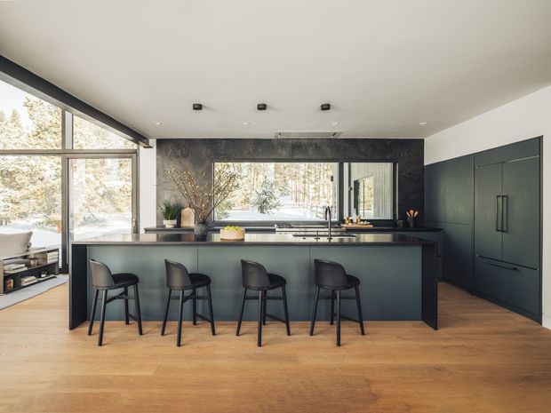 A contemporary kitchen with a long dark island and four bar stools, extensive integrated cabinetry along the right wall, and a dark stone or marble feature wall surrounding the cooktop and sink area. Large windows bring in natural light and overlook trees, while warm timber flooring contrasts with the charcoal cabinetry and surfaces.