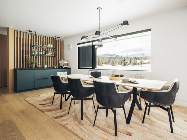 Bright contemporary dining room with a long light-wood dining table surrounded by dark upholstered chairs, set over a large patterned rug. A dark built-in sideboard and open barware shelving sit against a vertical timber-slat feature wall, while a large window provides expansive mountain views. A sculptural multi-arm pendant light hangs above the table.
