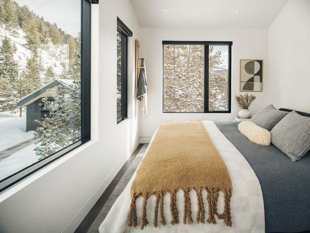A bright, compact bedroom with a low-profile bed positioned beside large black-framed windows overlooking a snowy forested landscape. The room has white walls, dark grey bedding, a cream and tan fringed throw, simple abstract wall art, and minimal natural decor. The clean-lined glazing and restrained furnishings create a calm, modern alpine retreat atmosphere.