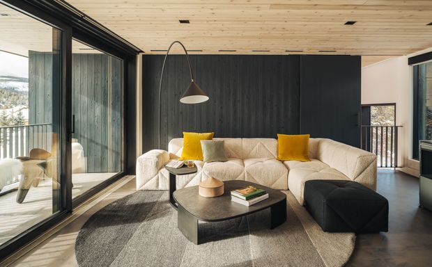 A warm, contemporary living room with a large cream sectional sofa, bright yellow accent cushions, black ottoman, and rounded dark coffee table. The room features extensive black-framed glazing opening to a balcony, a dramatic dark vertical timber feature wall, and a light timber-lined ceiling. A mountain landscape is visible outside.