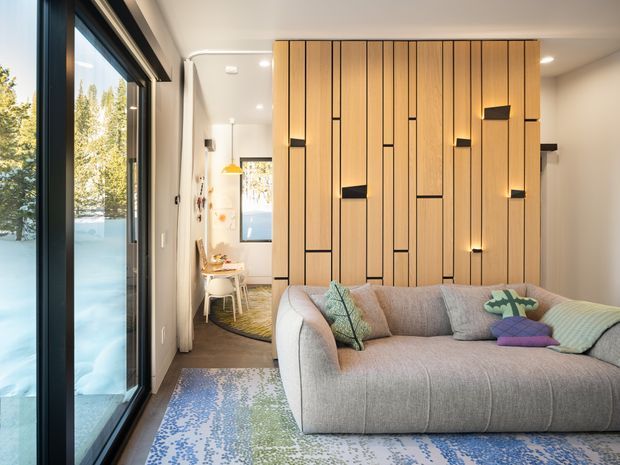 A bright contemporary living room with a large curved grey sectional sofa in the foreground, colourful patterned rug, and a full-height feature wall of vertical light timber battens with black recessed accents and integrated lighting. Black-framed floor-to-ceiling glazing opens to an outdoor view, while a small dining or workspace nook is visible at the rear.