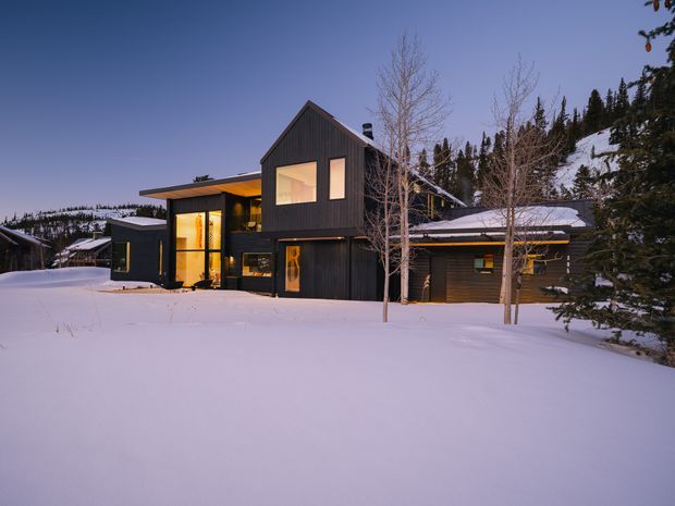 Exterior view of a contemporary dark-clad multi-level house set in a snowy alpine landscape at dusk. The building has steep pitched roof sections, large warm-lit windows, a projecting flat-roofed entrance volume, and an attached low garage-like wing. Snow blankets the foreground and surrounding terrain, with evergreen and leafless trees on the hillside behind.