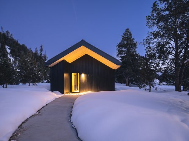 A small dark-clad cabin or outbuilding viewed from a snow-covered path at dusk. The compact single-storey structure has a prominent pitched gable roof with warm timber-toned soffits, vertical dark cladding, and a warmly illuminated glazed entry door. Deep snowbanks surround the walkway, with mature coniferous trees forming a wooded setting. The scene has a serene, warm contrast between the blue evening light, white snow, and amber entry lighting.