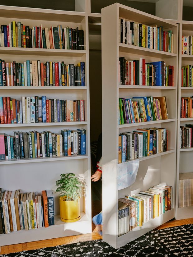 A bright home library or reading area with extensive white built-in floor-to-ceiling bookshelves filled with colourful books. A narrow central opening reveals part of a person and a hanging sheet, while a potted fern in a yellow planter sits on a lower shelf. Warm timber flooring and a black-and-white patterned rug add contrast.