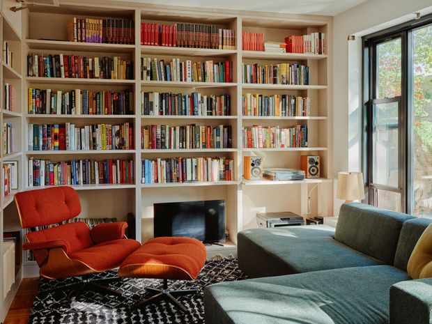A warm, book-filled living room with extensive built-in wall-to-wall shelving, a central television, and a compact media console. The room includes a red upholstered lounge chair with ottoman, a large blue-grey sectional sofa, a patterned dark rug, and tall black-framed windows bringing in natural light. The overall look is colourful, comfortable, and eclectic with mid-century-inspired furniture.
