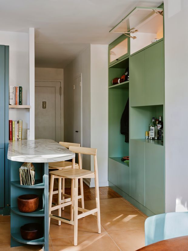 A compact, open-plan kitchen and entry area with a white marble island, integrated curved shelving, pale timber stools, and a full-height muted green built-in storage wall. The storage includes open shelving, concealed cabinetry, hanging rails, and a small drinks display. White walls, timber parquet flooring, and warm terracotta-toned floor tiles create a refined, colourful interior.