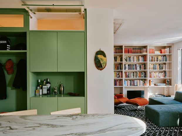 A stylish open-plan interior combining a kitchen or entertaining area with a living room and extensive built-in shelving. The foreground features a large rounded pale stone island or benchtop, while the left wall has sage-green built-in cabinetry with open storage, coat storage and a drinks display. Beyond the dividing wall is a library-lined living area with colourful lounge chairs, a deep teal sofa and a patterned dark rug. The space has white walls and ceilings, warm timber shelving, and a richly layered, eclectic character.