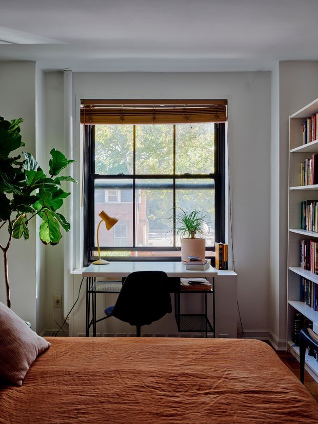 A compact, bright bedroom with a built-in desk positioned in front of a large multi-pane window. The room includes a bed with a rust-colored cover, a black desk chair, a yellow desk lamp, potted plants, open wall shelving filled with books, and warm wooden window blinds. White walls and trim create a simple, functional interior.