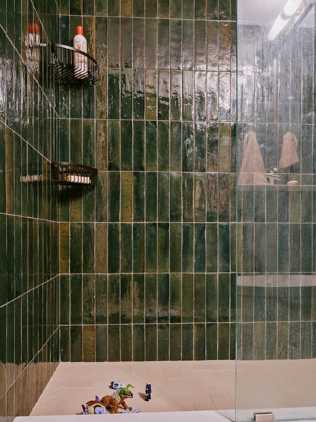 Close-up view of a compact shower area with vertically arranged glossy deep green ceramic tiles, pale grout, black wire shower caddies, and a clear glass shower screen. A reflected showerhead and bath toys are visible near the base, adding a lived-in detail.