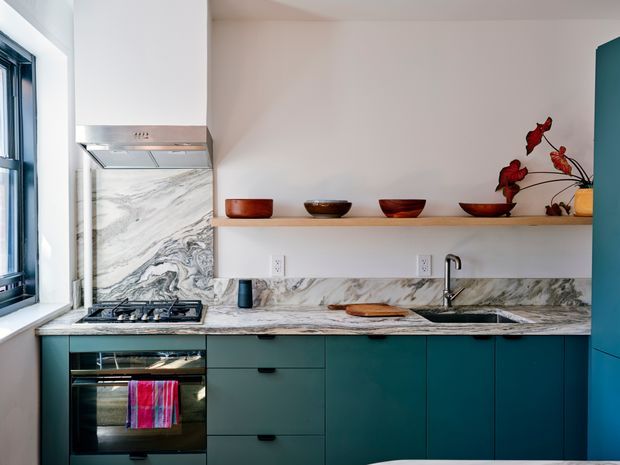 A compact contemporary kitchen with deep teal flat-panel cabinetry, a pale marble-look stone benchtop and splashback, integrated gas cooktop and oven, stainless steel rangehood, and a single-bowl sink. A floating timber shelf displays wooden bowls and decorative objects, while a large window brings in natural light.