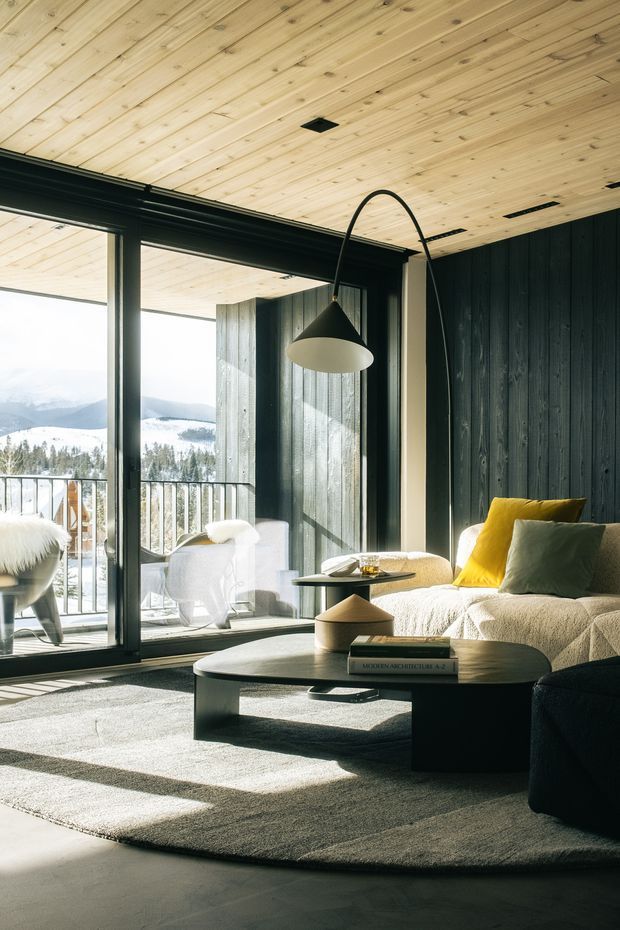 A warm, modern living room with expansive black-framed glazing opening to a balcony and mountain landscape. The room features a pale timber-lined ceiling, dark vertical timber wall panelling, a light upholstered sofa with mustard and sage cushions, a low oval coffee table, a sculptural arched floor lamp, and a large neutral rug. Strong sunlight creates dramatic shadows across the floor and furnishings.