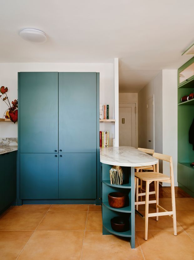 A compact, brightly styled kitchen and dining nook with deep teal built-in cabinetry, a curved marble-look benchtop forming a small peninsula, open shelving, timber stools, and terracotta tile flooring. A narrow hallway and additional green storage joinery are visible beyond the main kitchen area.