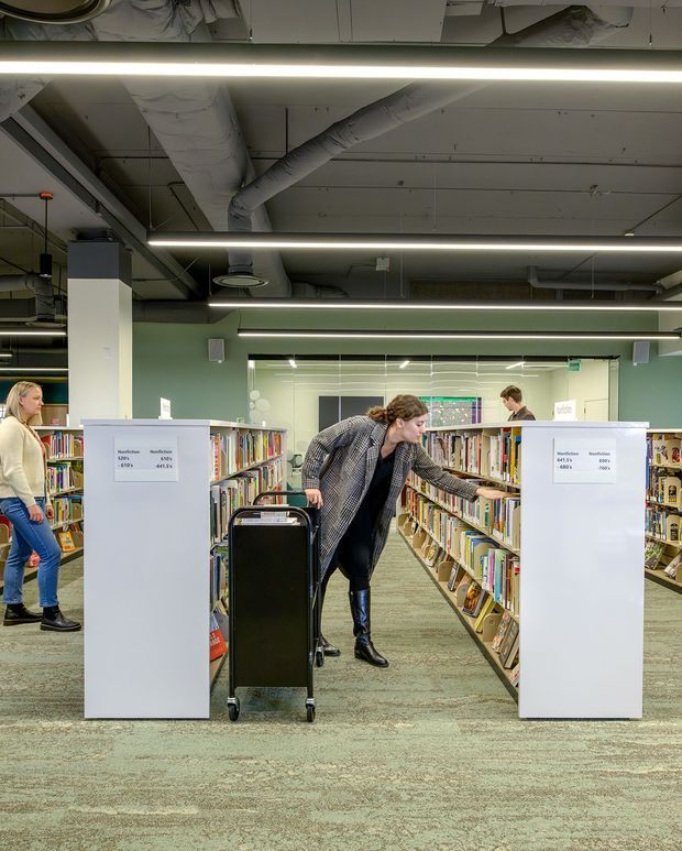 A contemporary public library interior viewed down rows of bookshelves, with patrons browsing and a library cart in the foreground. The space has muted sage-green walls, carpeted flooring, white shelving and partitions, exposed services and ductwork at the ceiling, and long linear fluorescent-style strip lighting. A glazed interior wall or reflection appears at the rear, contributing to the bright, functional commercial setting.