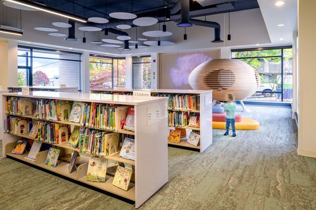 A bright contemporary children's library or reading area with low open bookshelves filled with picture books, a central circulation/storage unit, carpeted flooring, extensive windows, and a distinctive suspended oval timber reading pod. The exposed dark ceiling services and playful acoustic ceiling discs contrast with light joinery and colourful children's furnishings.