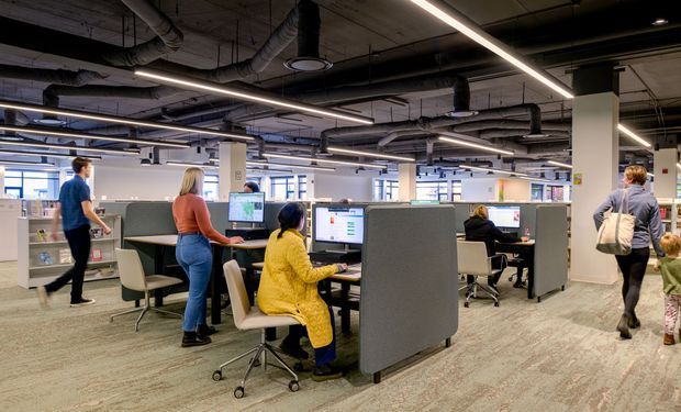 Open-plan commercial office interior with rows of workstation pods, grey acoustic privacy screens, computer monitors, rolling office chairs, exposed ceiling services and ducts, and linear strip lighting. Several people are working or moving through the space. The office has a spacious, bright, contemporary workplace fit-out with a carpeted floor and large windows.