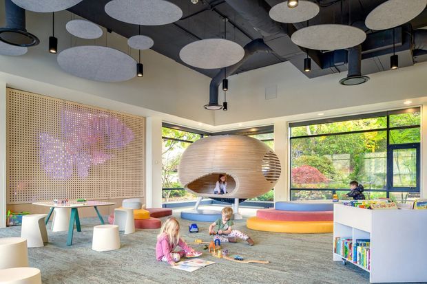 Bright, playful contemporary children's learning and activity room with large windows, low colourful seating, children’s furniture, bookshelves, and a distinctive suspended timber pod structure. The ceiling has exposed dark services with numerous acoustic discs and pendant lights; a perforated timber feature wall adds texture.