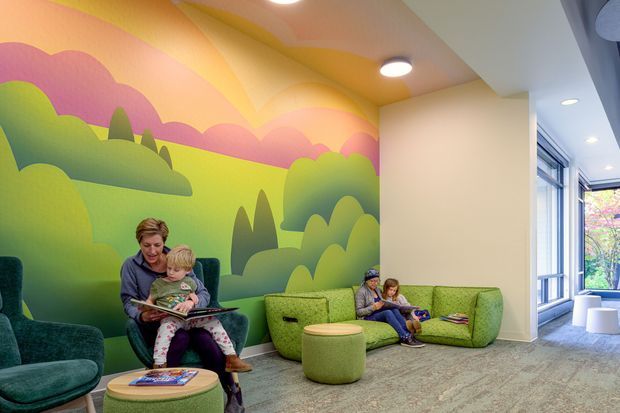 A bright, playful children's reading and waiting area with green upholstered lounge chairs and sofas, low round ottomans, and books. A large colourful mural of rolling hills, trees, and clouds covers the back wall and continues across the ceiling. Floor-to-ceiling glazing and a corridor or adjoining space are visible on the right.