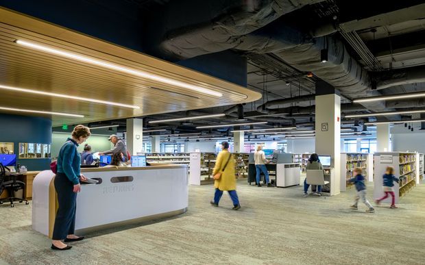 An expansive contemporary public library interior with an information or reception desk in the foreground, open-plan study areas, bookshelves, computers, and people using the space. The ceiling exposes extensive mechanical services and ductwork, contrasted with a suspended curved timber-look slatted canopy and integrated linear lighting above the desk. The palette is predominantly cool grey, white, and blue with warm wood accents and carpeted flooring.