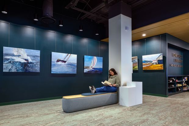 A contemporary dark-toned gallery or library interior with deep blue-green paneled walls, multiple framed artworks, integrated display shelving, and a built-in upholstered bench. A visitor is seated reading near a sculptural white column and pedestal. The ceiling has exposed services and recessed spot lighting, while a warm timber slatted ceiling section adds contrast.