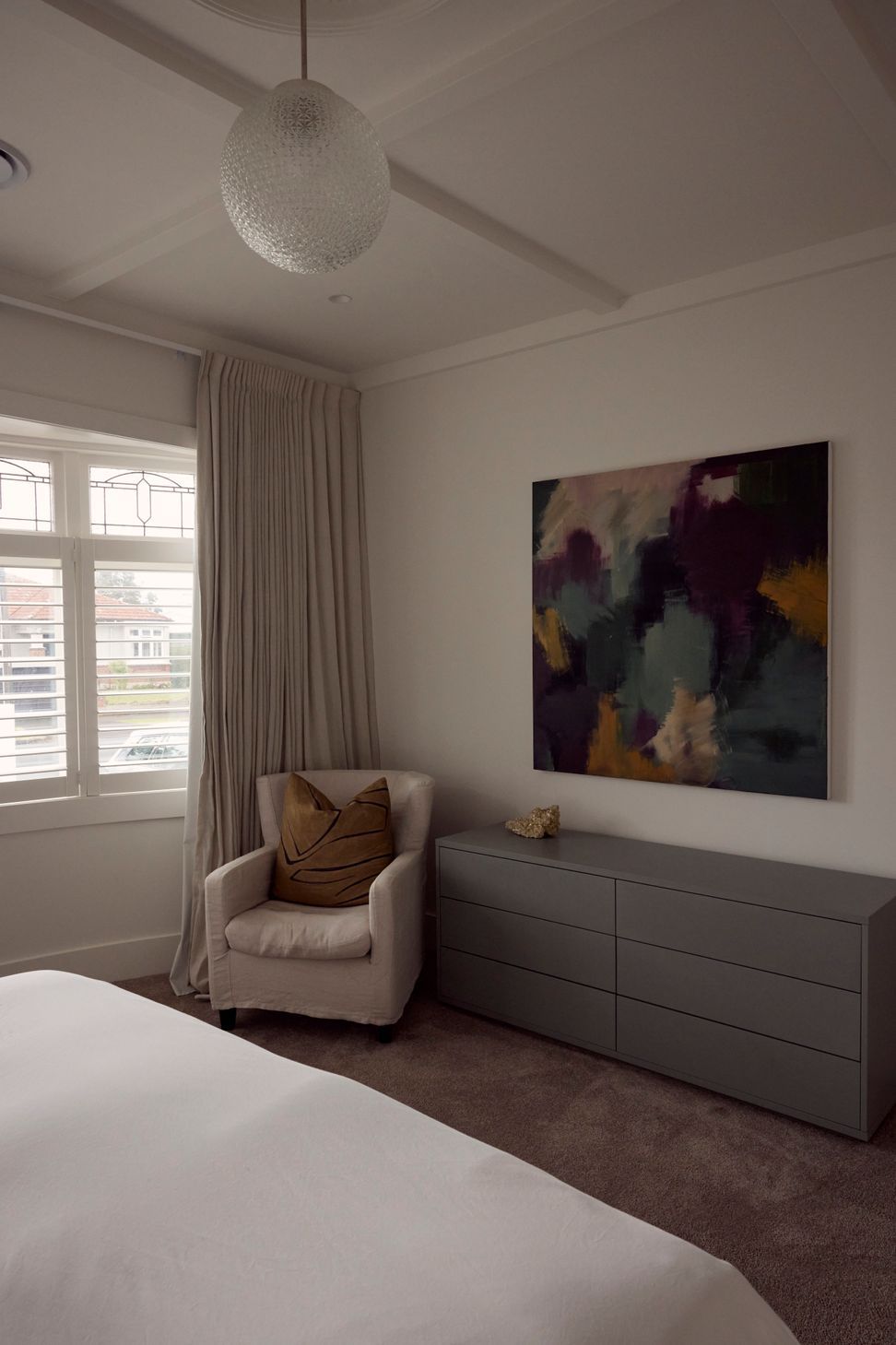 Large format abstract artwork above a grey chest of drawers, linen armchair and original leadlight windows — the bedroom at Point Chevalier Bungalow. In collaboration with Hannah Warren Interiors.