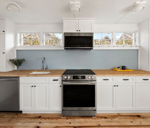 Bright, compact kitchen with white Shaker-style cabinetry, a stainless-steel range and dishwasher, pale blue subway-tile splashback, timber benchtops, and horizontal windows above the work area. The space has a clean, classic coastal feel with warm timber flooring and simple black hardware.