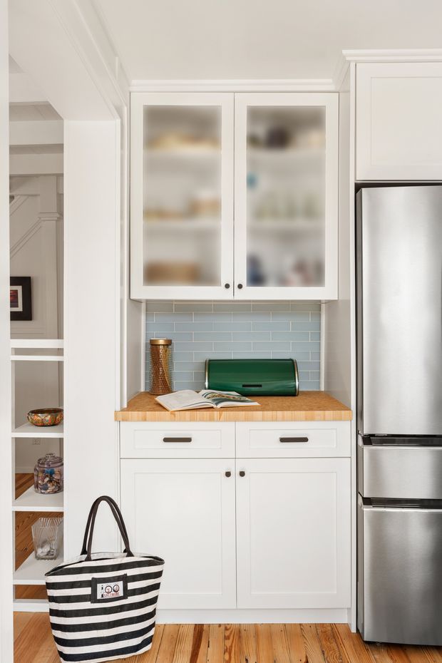 A compact, neatly fitted kitchen pantry or appliance nook with white Shaker-style cabinetry, frosted-glass upper doors, a pale blue subway-tile splashback, timber benchtop, stainless-steel refrigerator, and warm timber flooring. Open shelving is visible to the left, with small kitchen items and a striped tote bag in the foreground. The space has a bright, clean, traditional character with restrained coastal styling.