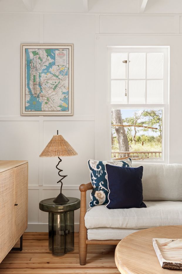 A bright, cozy living room with white paneled walls, a natural timber floor, and a large traditional sash window overlooking trees. A light upholstered sofa with navy and patterned cushions sits beside a woven rattan-shaded table lamp and a dark glass side table. A timber cabinet, framed map artwork, and part of a round timber coffee table complete the relaxed coastal-inspired interior.