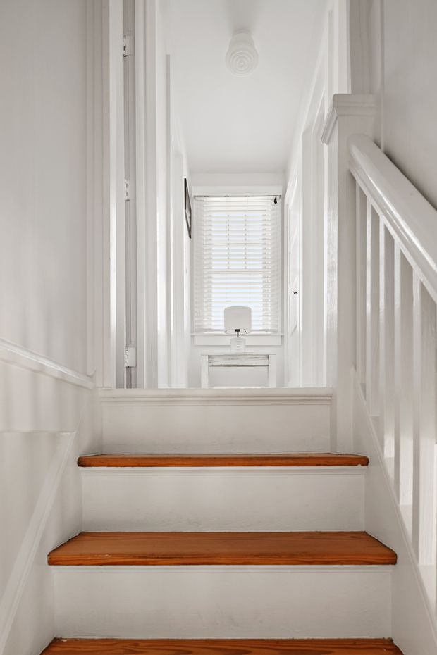 Bright, narrow residential stairway leading up to a small landing or hallway. The staircase has white-painted risers, walls, balustrade and trim, with warm stained timber stair treads. At the top is a white console table with a lamp beneath a window fitted with white horizontal blinds. The space has traditional detailing and a clean, monochromatic appearance.