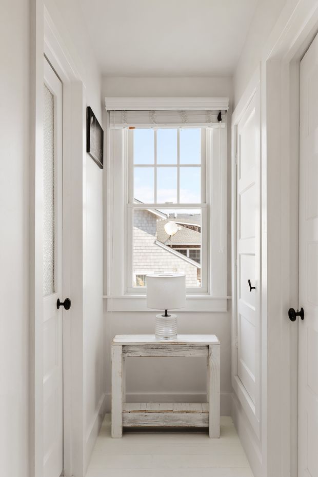 Bright, narrow white hallway or landing with paneled doors on both sides, a large multi-pane window at the end, and a small distressed white console table topped with a ribbed table lamp. White trim, walls, ceiling, and light flooring create a clean, airy setting; rooftops are visible through the window.